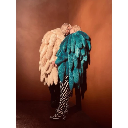 Two people in large feather coats stand close together in a corner with an orange wall, one wearing zebra print boots.