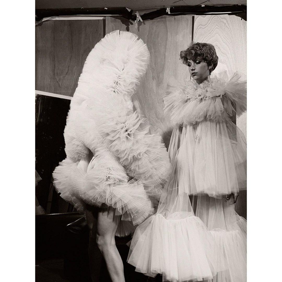 Black and white photograph of two models wearing elaborate white tulle gowns, one partially obscured, posing in front of a wooden background.
