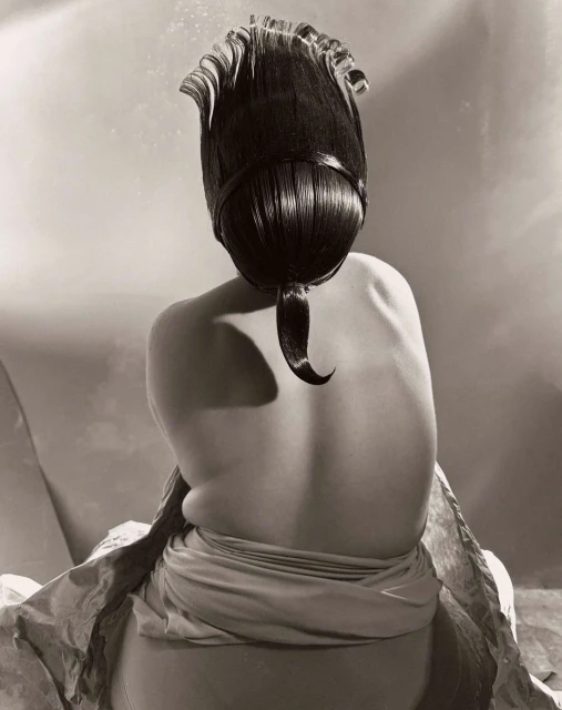 Back view of a person with an elaborate dark hairstyle, wearing a draped garment, in a monochromatic artistic studio photograph.