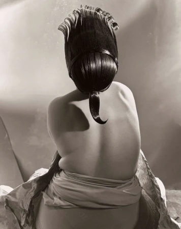 Back view of a person with an elaborate dark hairstyle, wearing a draped garment, in a monochromatic artistic studio photograph.