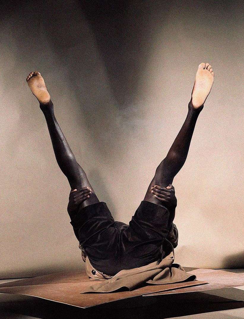 Adult figure lying on back, legs extended, hands holding thighs in indoor studio, against neutral gradient background in creative art photography.
