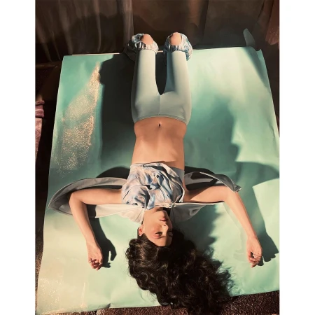 Woman lying on back on blue surface with head at bottom and legs extending upward; wearing blue leggings, shirt, and foot coverings.