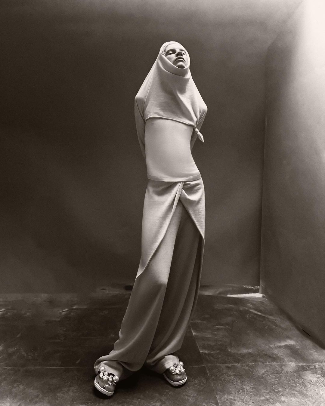 Monochromatic fashion portrait of a person in draped clothing with a head covering, posing in front of a studio backdrop.
