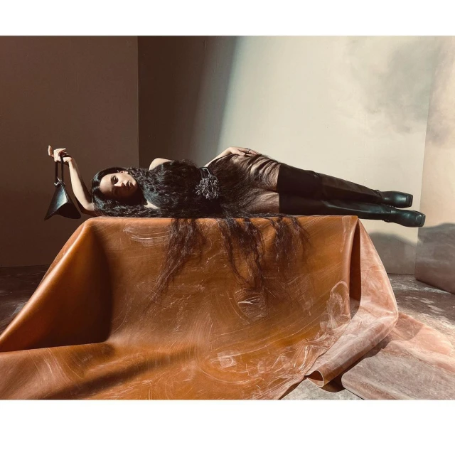 Woman in a dark sheer dress and boots, lying on a brown surface, holding a triangular purse. Dark hair spread around her. Fashion editorial.