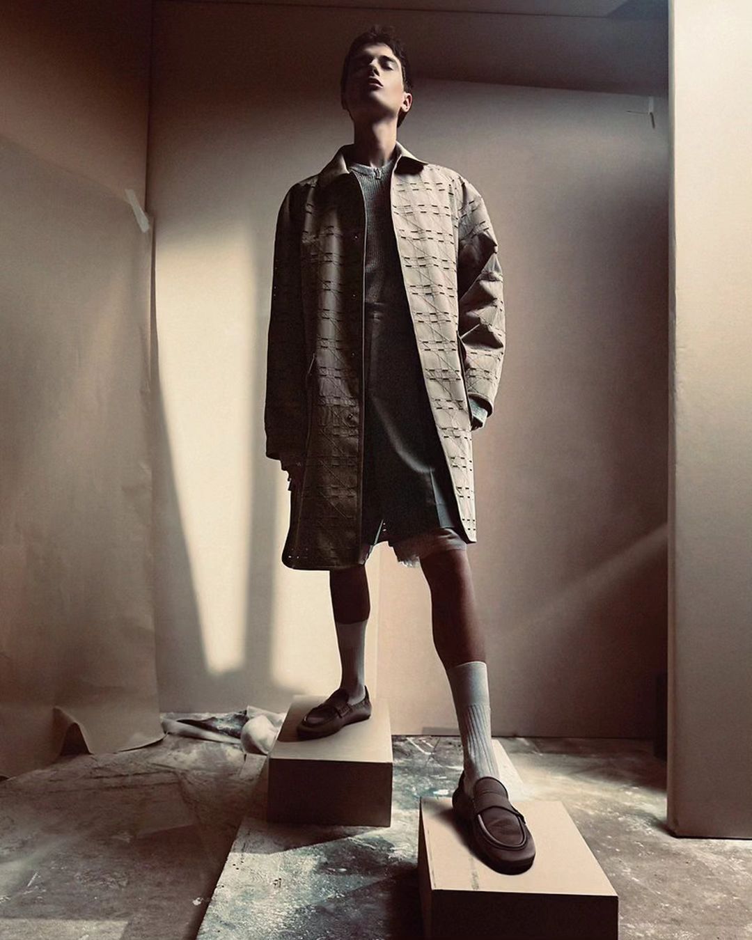 A young adult male model poses on blocks in a studio wearing a patterned brown coat, grey top and shorts, white socks, and brown loafers.
