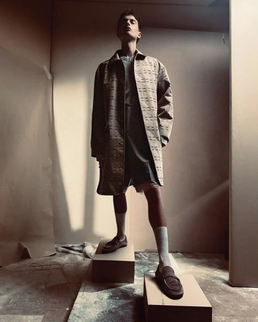 A young adult male model poses on blocks in a studio wearing a patterned brown coat, grey top and shorts, white socks, and brown loafers.