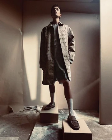 A young adult male model poses on blocks in a studio wearing a patterned brown coat, grey top and shorts, white socks, and brown loafers.