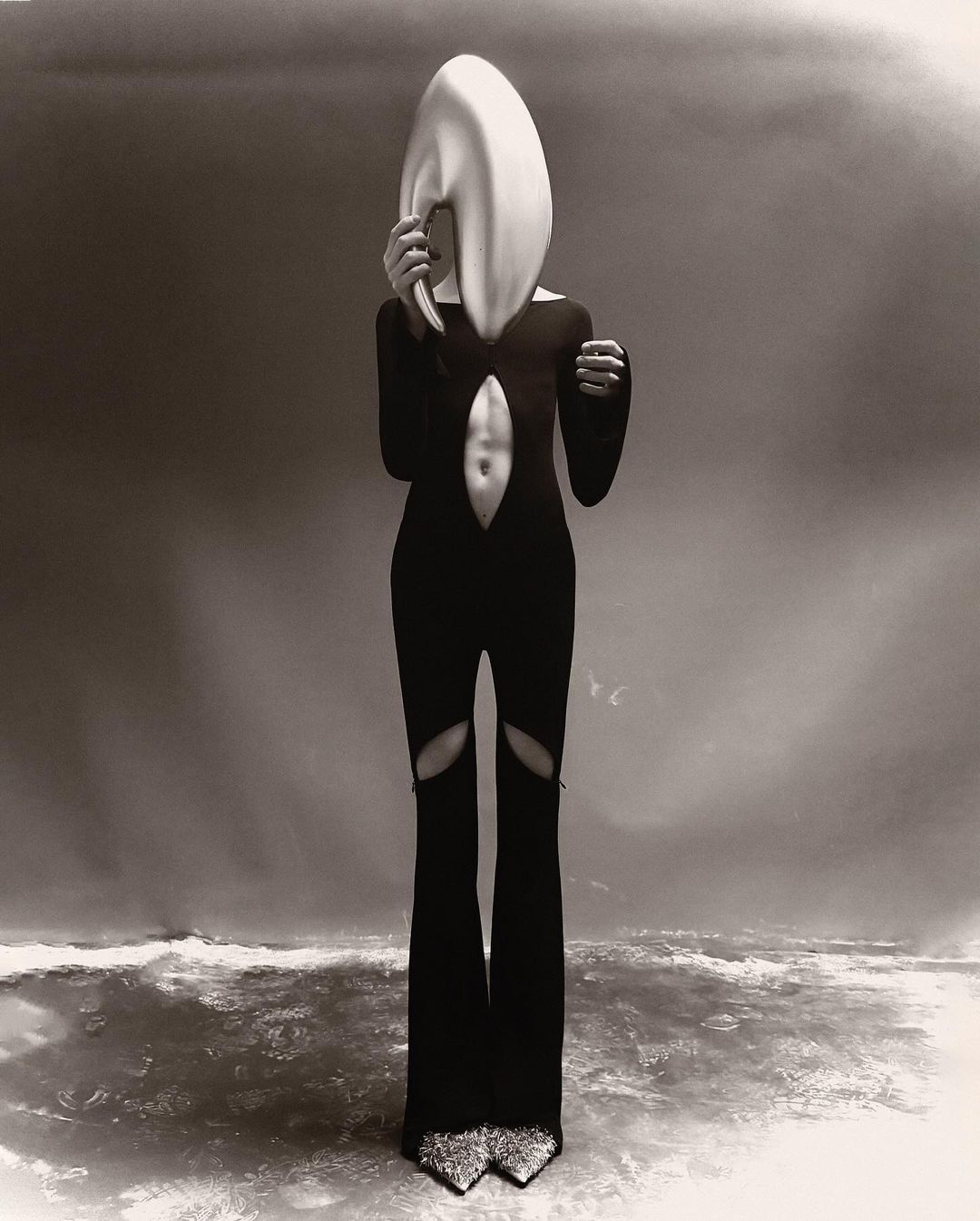 A person in a black bodysuit and statement headpiece poses in front of a gradient background, creating a surreal fashion editorial.