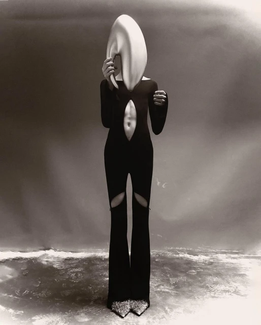 A person in a black bodysuit and statement headpiece poses in front of a gradient background, creating a surreal fashion editorial.