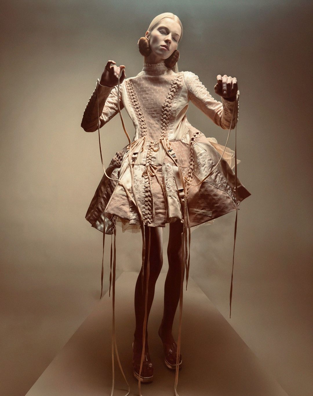 A slender, fair-skinned model in a pale pink, marionette-inspired dress poses in a photo studio with controlled strings creating a dramatic effect.