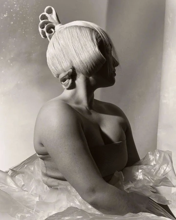 Monochrome portrait of an adult female with an elaborate blonde hairstyle and pale skin, posing in a photo studio with neutral background.