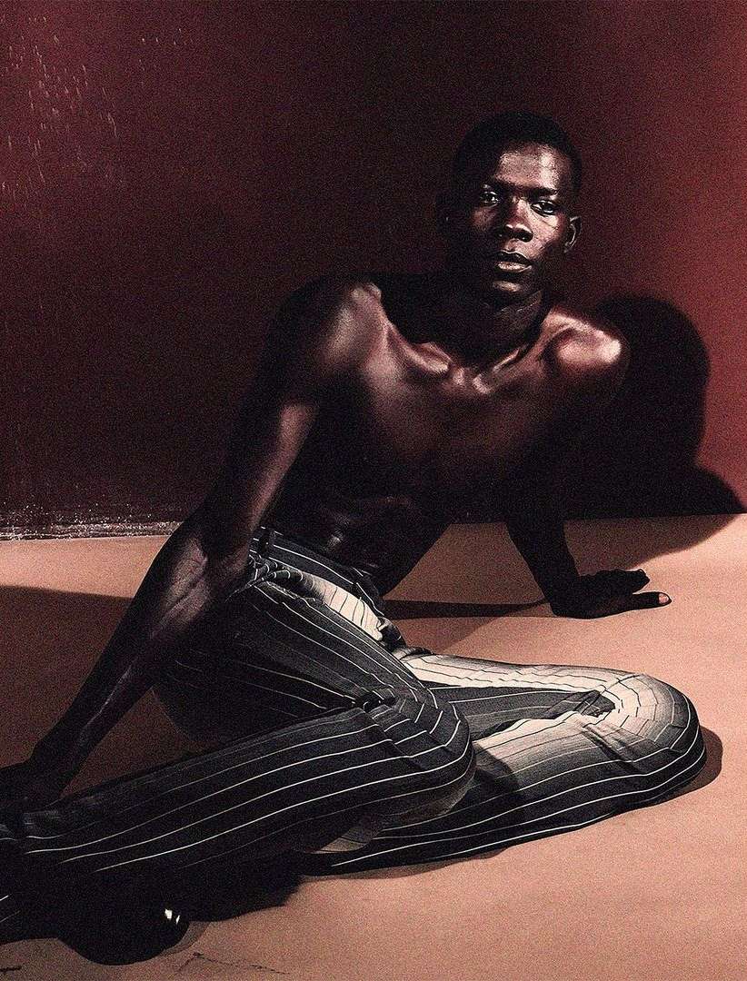 Portrait of a shirtless African American male sitting on the floor wearing striped pants in a studio with red wall.