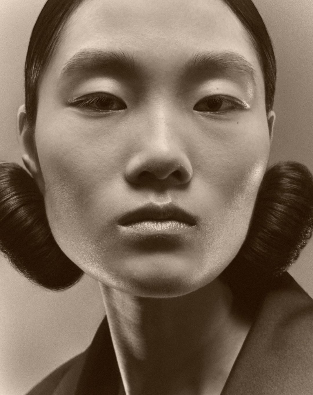Close-up portrait of a woman with styled hair and minimalist makeup, in a sepia tone studio setting, showcasing beauty and fashion.