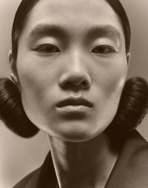 Close-up portrait of a woman with styled hair and minimalist makeup, in a sepia tone studio setting, showcasing beauty and fashion.