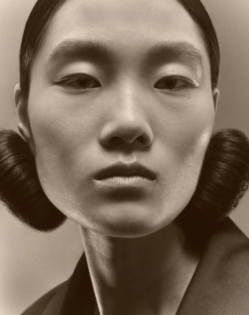 Close-up portrait of a woman with styled hair and minimalist makeup, in a sepia tone studio setting, showcasing beauty and fashion.