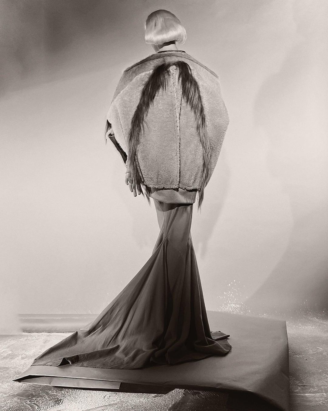 Model seen from behind wearing an elaborate structured jacket with dark wing-like elements and a long flowing skirt on a studio backdrop.