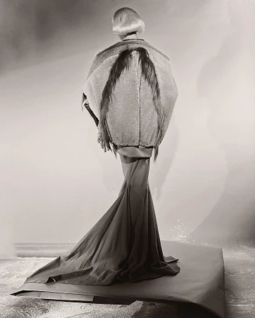 Model seen from behind wearing an elaborate structured jacket with dark wing-like elements and a long flowing skirt on a studio backdrop.