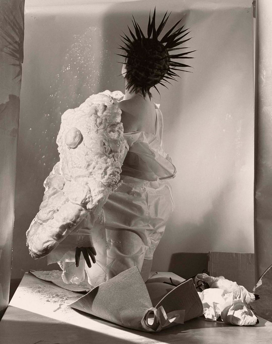Avant-garde fashion photo: A person in an artful white garment and spiky hair faces away, showcasing unique textures and design in studio setting.