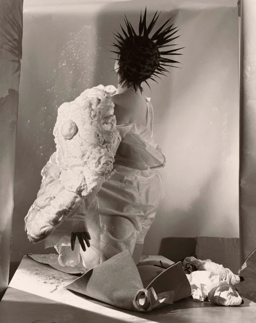 Avant-garde fashion photo: A person in an artful white garment and spiky hair faces away, showcasing unique textures and design in studio setting.