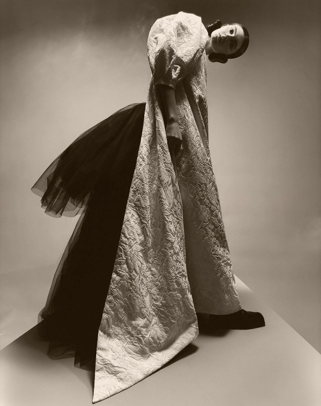 Fashionable woman poses wearing long white textured dress with black tulle accents in studio with soft sepia lighting. 