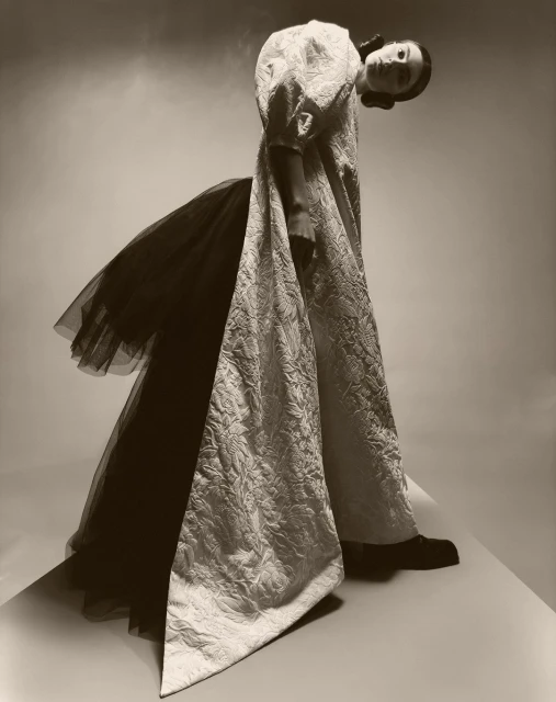 Fashionable woman poses wearing long white textured dress with black tulle accents in studio with soft sepia lighting. 