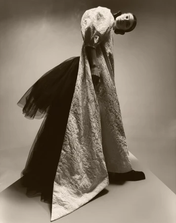 Fashionable woman poses wearing long white textured dress with black tulle accents in studio with soft sepia lighting. 
