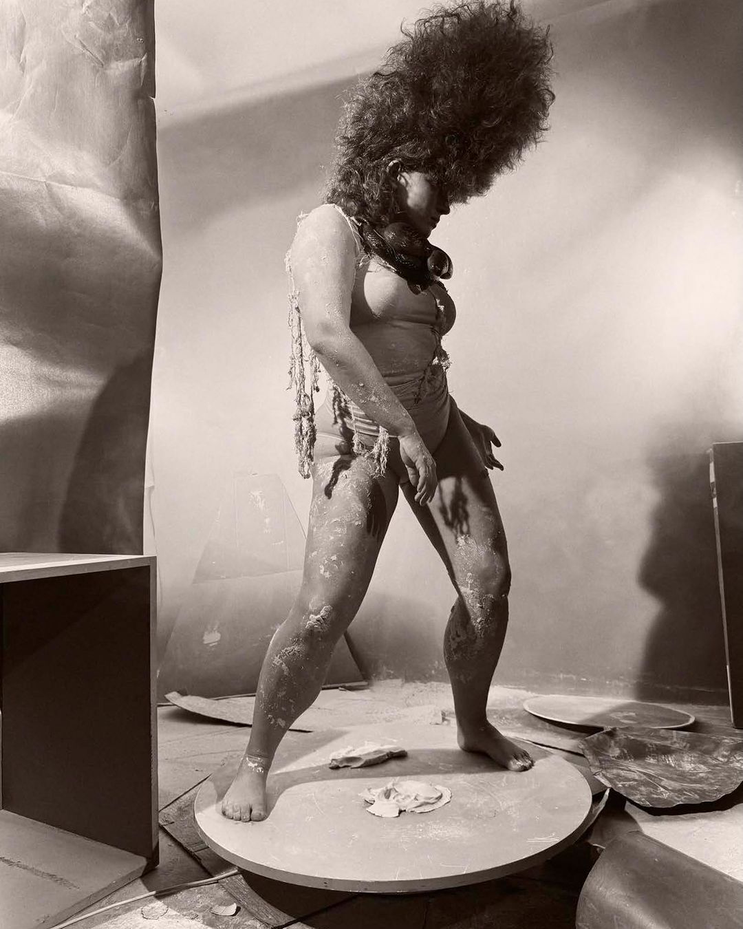 Sepia-toned photo of person covered in powder on platform, wearing light-colored swimsuit and beads, against draped background in studio.