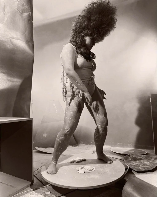 Sepia-toned photo of person covered in powder on platform, wearing light-colored swimsuit and beads, against draped background in studio.