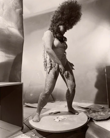 Sepia-toned photo of person covered in powder on platform, wearing light-colored swimsuit and beads, against draped background in studio.