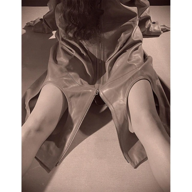 An adult female lies on a beige surface wearing a light brown leather coat, hair visible near the shoulders. The coat is partially open.