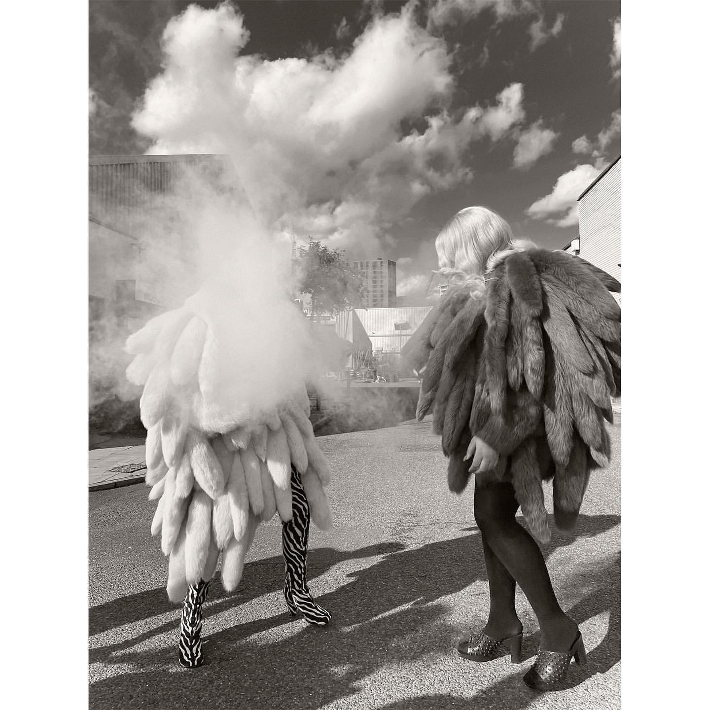 Monochrome outdoor fashion shot of two figures wearing elaborate fluffy clothing on a city street on a cloudy day.