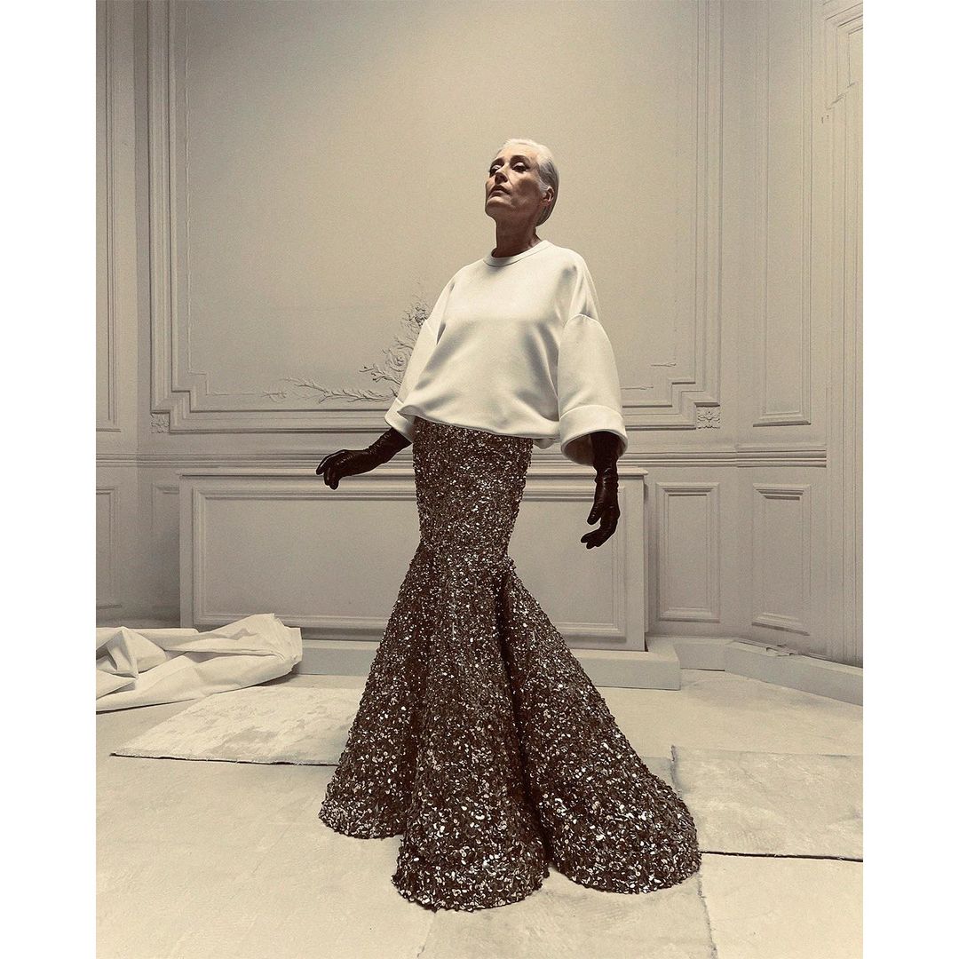Elegant older woman poses in a white top and shimmering gold fishtail skirt in a photo studio with paneled walls and soft, diffused lighting.