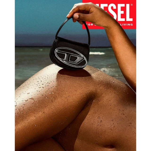 Black Diesel handbag with a silver logo rests on a wet tan surface, potentially skin, against a blurry background of beach and ocean.