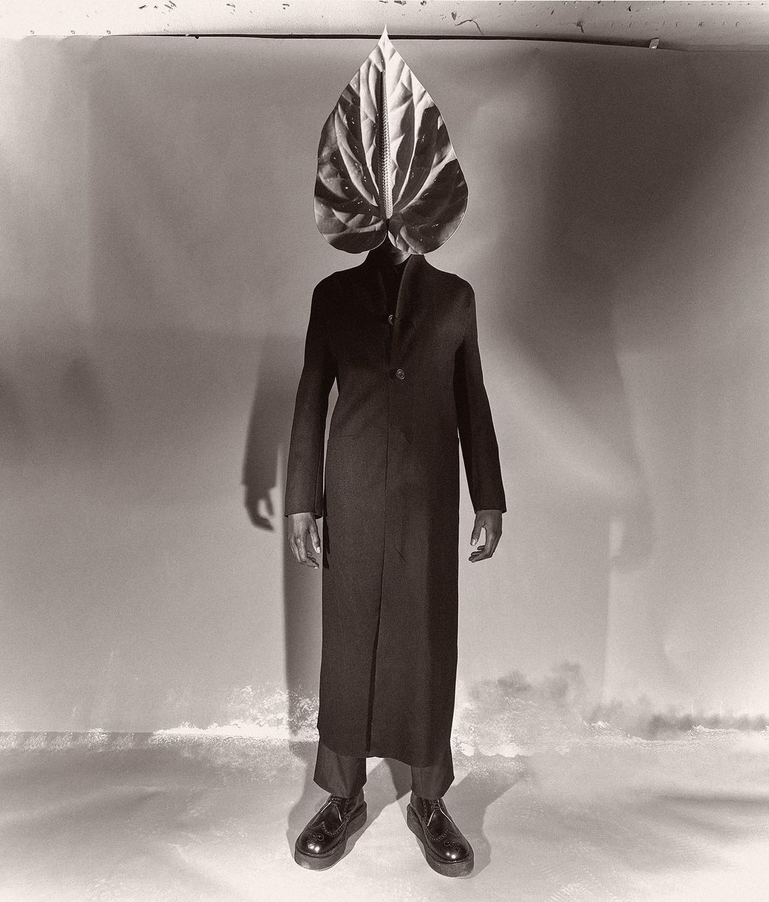 Full-length studio portrait of a person with a large Anthurium leaf over their head, wearing a dark coat and shoes against a mottled backdrop.