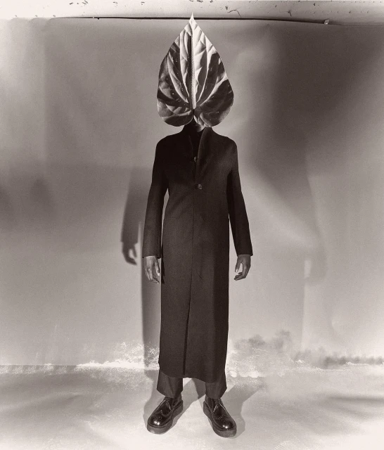 Full-length studio portrait of a person with a large Anthurium leaf over their head, wearing a dark coat and shoes against a mottled backdrop.