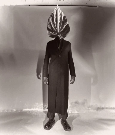Full-length studio portrait of a person with a large Anthurium leaf over their head, wearing a dark coat and shoes against a mottled backdrop.