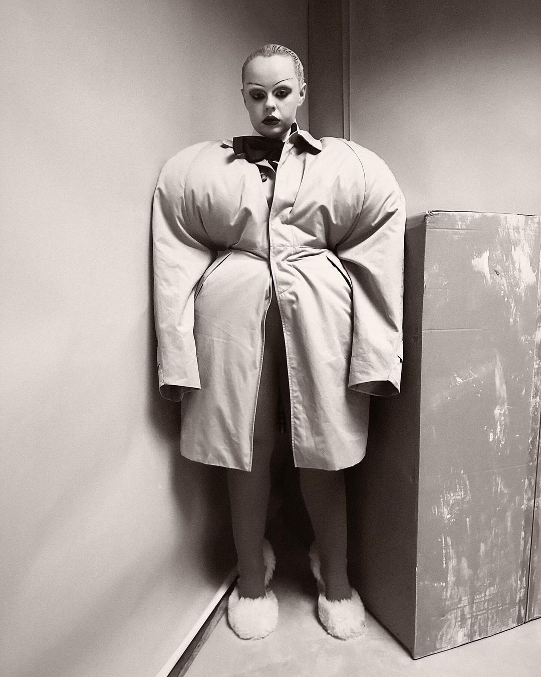 Monochrome portrait of a woman in an avant-garde, oversized beige trench coat with voluminous sleeves, and fluffy slippers, posing indoors.