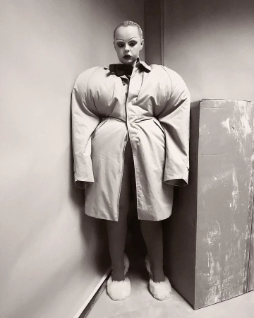 Monochrome portrait of a woman in an avant-garde, oversized beige trench coat with voluminous sleeves, and fluffy slippers, posing indoors.