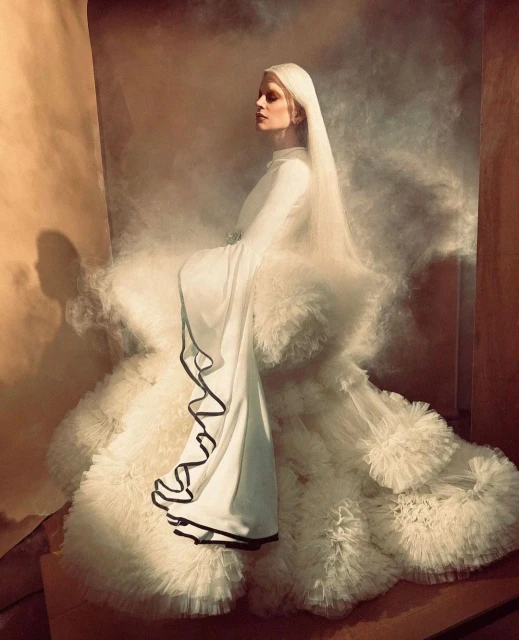 Elegant woman in white gown with veil poses against a smoky, neutral backdrop in a fashion editorial photo shoot, side view.