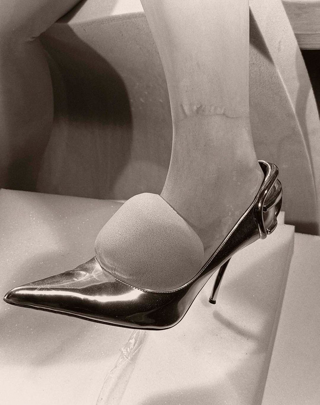 Close-up on foot wearing shiny stiletto heel pump, with toe insert, on white textured surface, in monochrome. Fashionable footwear.