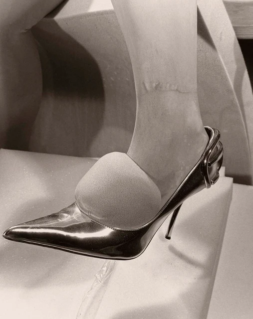 Close-up on foot wearing shiny stiletto heel pump, with toe insert, on white textured surface, in monochrome. Fashionable footwear.