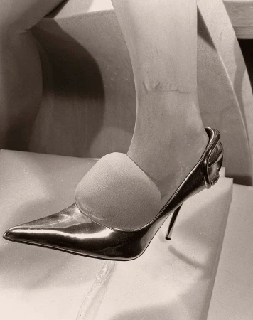Close-up on foot wearing shiny stiletto heel pump, with toe insert, on white textured surface, in monochrome. Fashionable footwear.