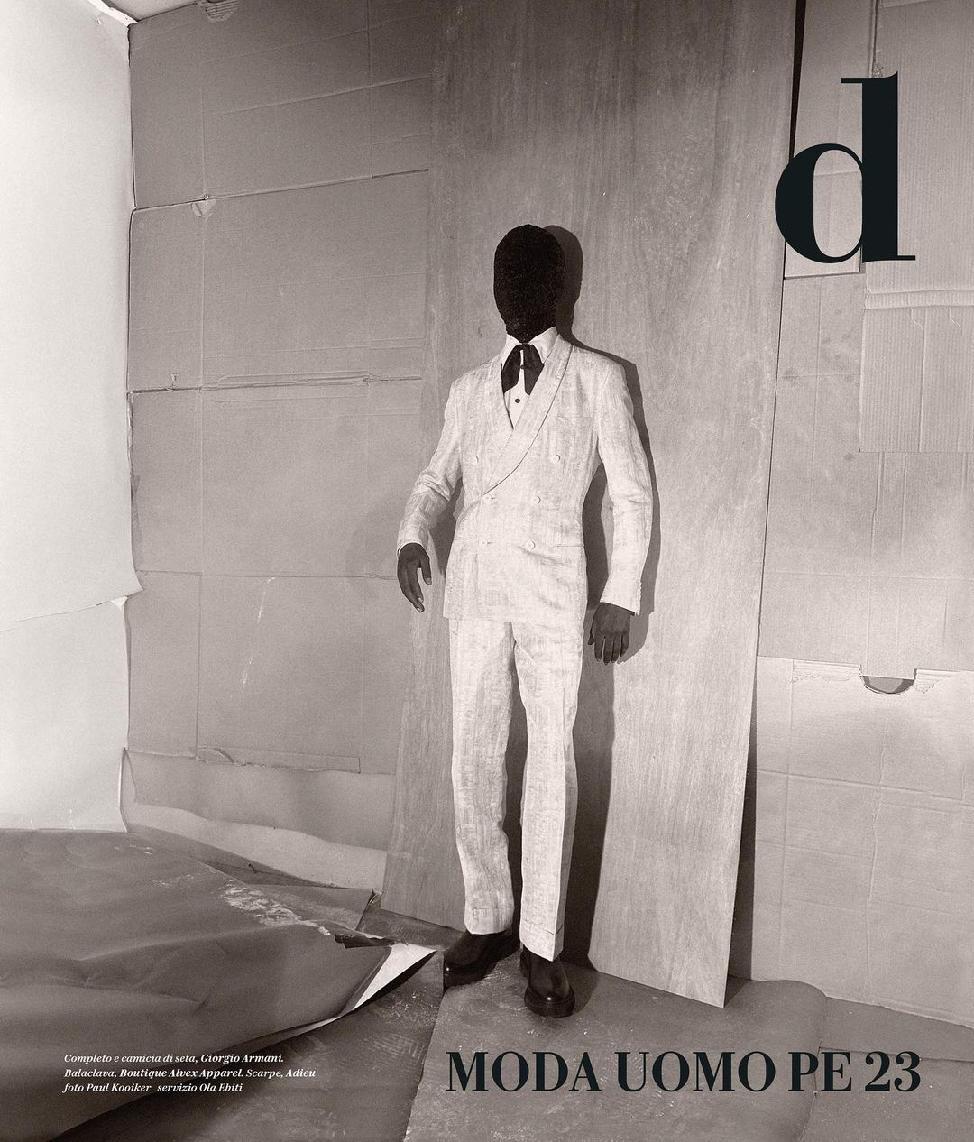 Monochrome fashion shot of a faceless person in an ivory suit, standing against a layered cardboard background with bold black text overlay.