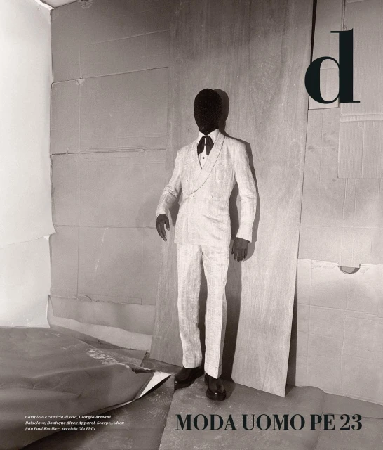 Monochrome fashion shot of a faceless person in an ivory suit, standing against a layered cardboard background with bold black text overlay.