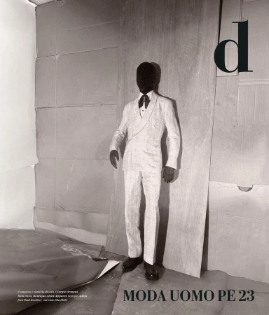 Monochrome fashion shot of a faceless person in an ivory suit, standing against a layered cardboard background with bold black text overlay.