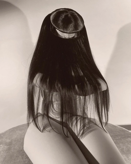 A person with long black hair draped over their body is seated in an indoor setting, against a neutral background in a monochromatic style.