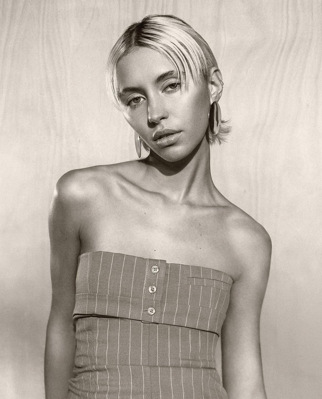 Portrait of a young adult woman with short blonde hair wearing a pinstripe tube top, posing against a neutral wood-panel backdrop.