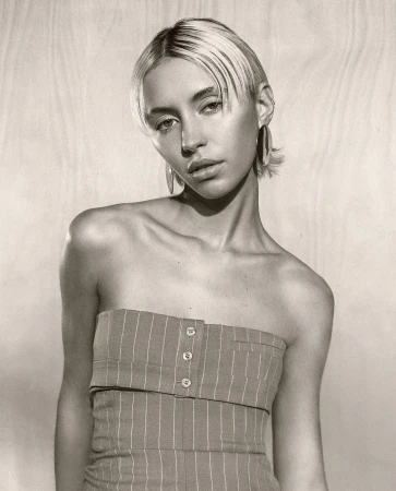 Portrait of a young adult woman with short blonde hair wearing a pinstripe tube top, posing against a neutral wood-panel backdrop.