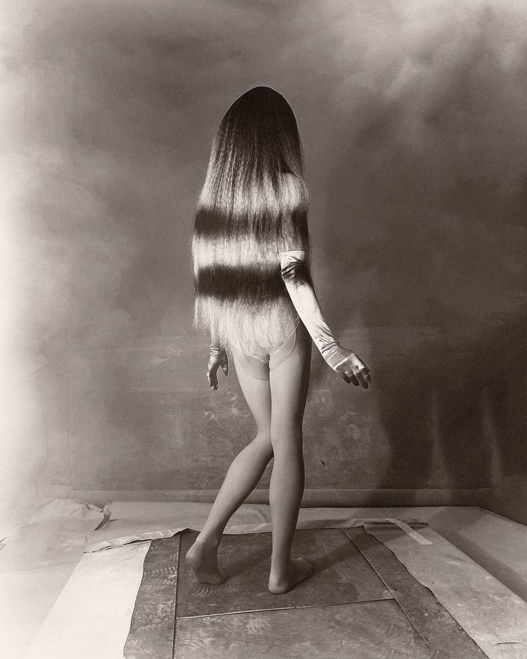 Monochrome photograph of a person with long, horizontally striped hair wearing gloves and minimal clothing posing in a studio environment.