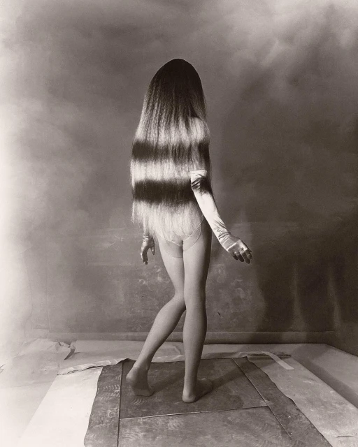 Monochrome photograph of a person with long, horizontally striped hair wearing gloves and minimal clothing posing in a studio environment.